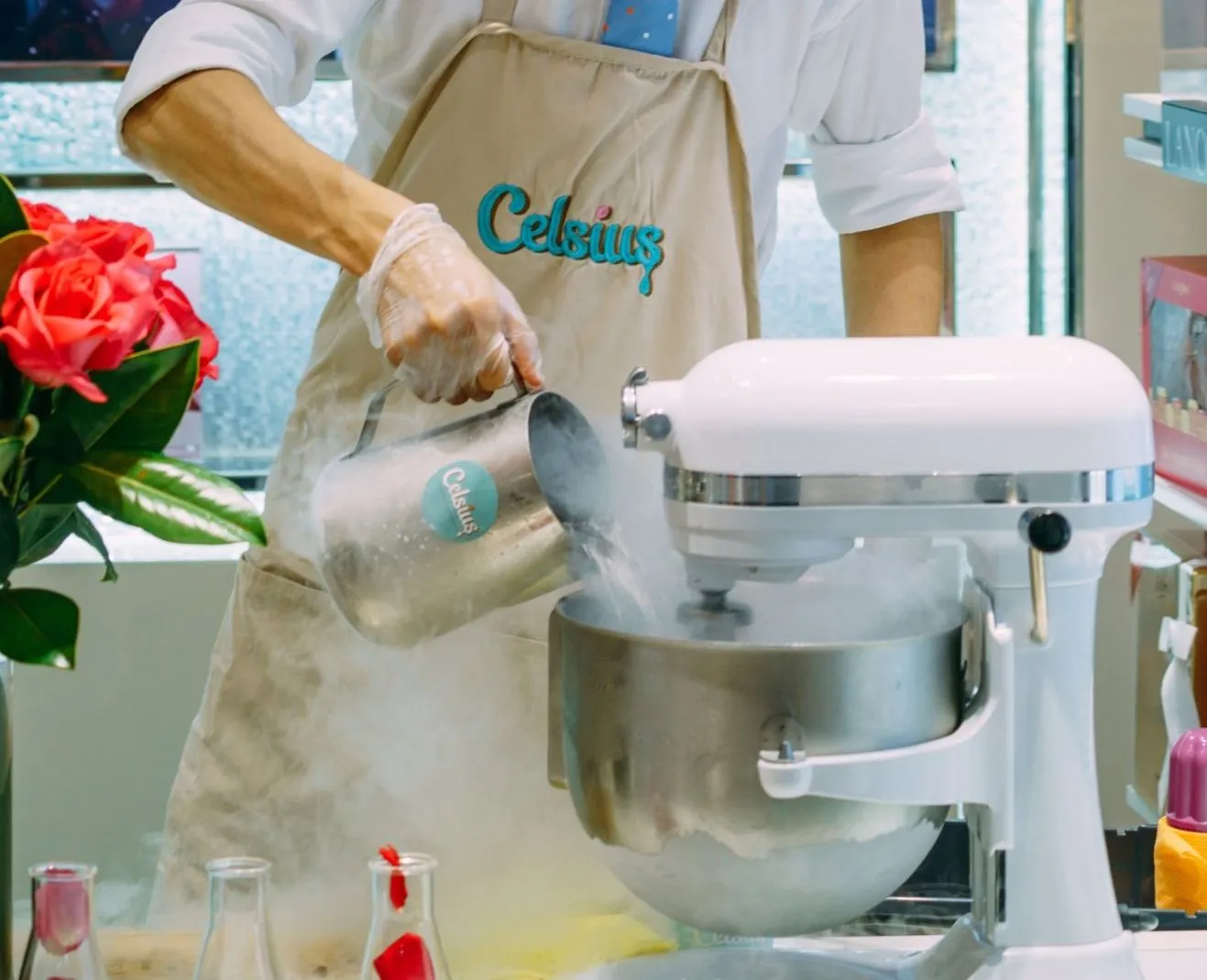 A gloved hand pours liquid nitrogen from a steel jug into a mixer bowl, fog spilling over the rim, beside a vase of roses